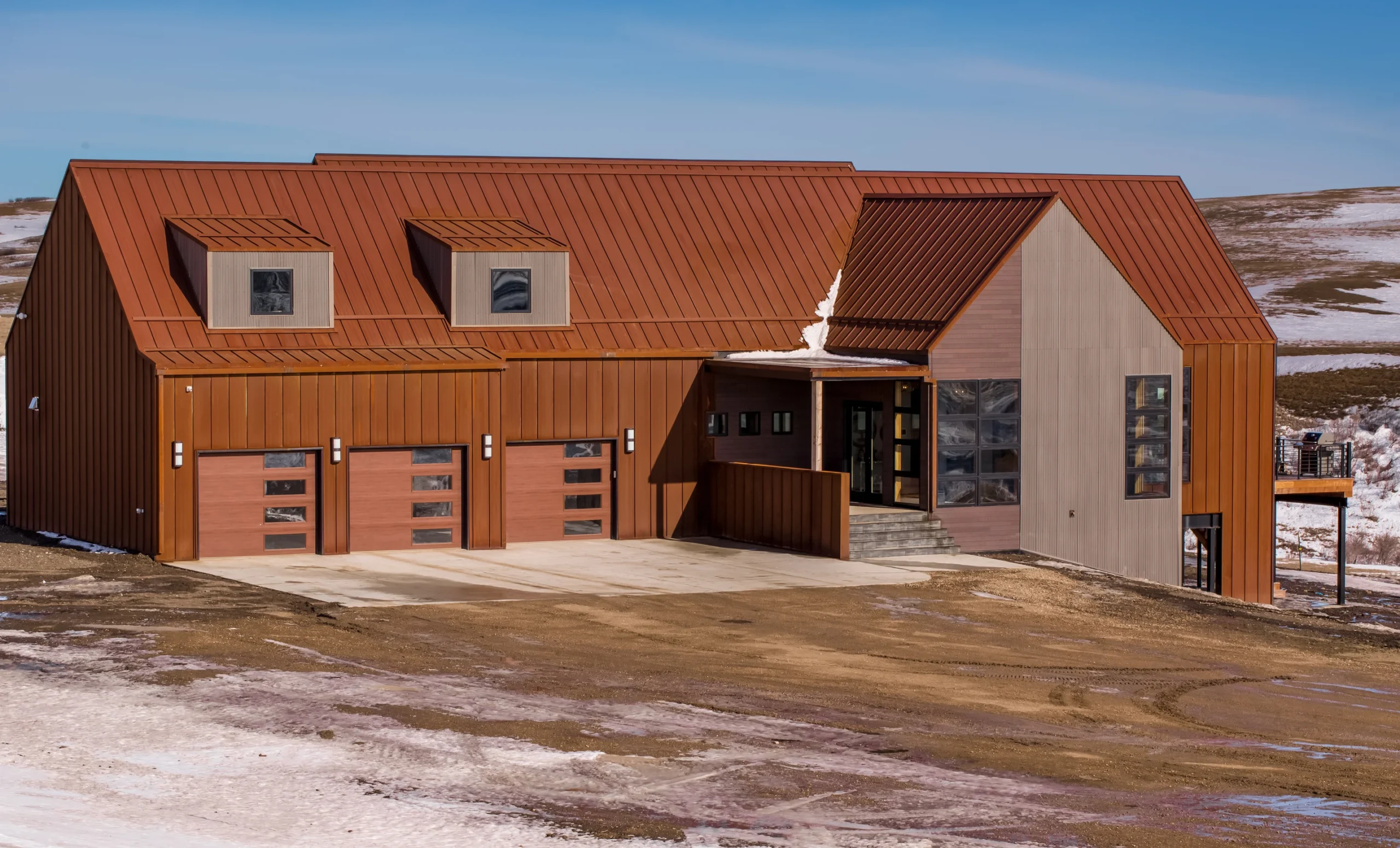 exterior view of large home of SIP construction; brown siding and roofing; three-car garage; unique windows; setting is windswept prarier with light snow, muddy tire tracks in the fore - photo