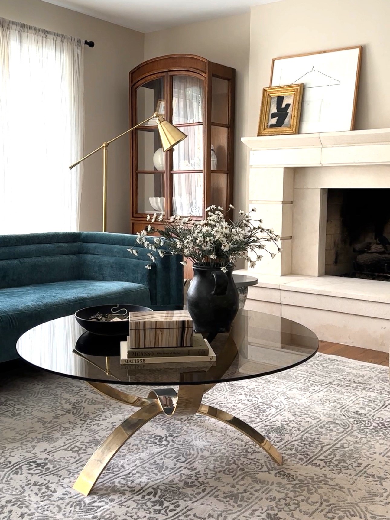 lovely interior home, vintage brass and glass round coffee table and colorful modern couch; bright window, fireplace, and wooden armoire - photo